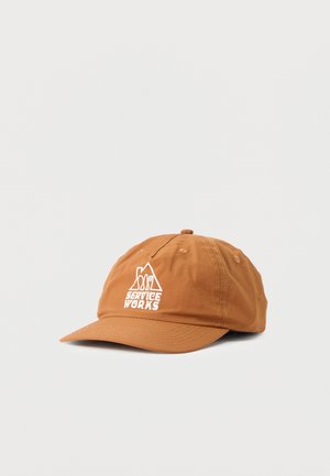 Casquette de baseball marron clair avec le logo blanc "Service Works" et un dessin de montagne à l'avant, sur un fond gris uni.