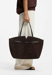 Henkilö pitää kädessään suurta tummanruskeaa mokkanahkaisesta materiaalista valmistettua olkalaukkua, jossa on mustat kahvat. Hänellä on päällään kermaiset housut ja ruskea neulepaita, ja hän seisoo valkoista taustaa vasten.