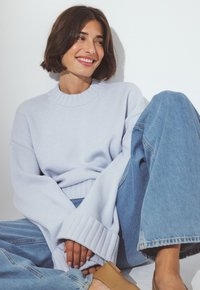 Leende kvinna med kort brunt hår som bär en ljusblå oversized tröja och lösa blå jeans, sitter avslappnat mot en vit vägg.