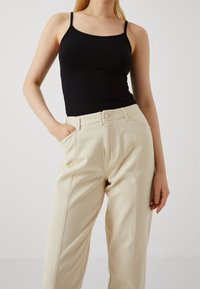 Débardeur noir ajusté à fines bretelles, associé à un pantalon taille haute beige clair avec poches latérales et détails de coutures visibles.
