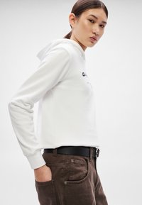 Hanorac alb scurt, cu mâneci lungi, cu un logo imprimat negru. Asortat cu pantaloni maro și o curea neagră cu detaliu de cataramă.