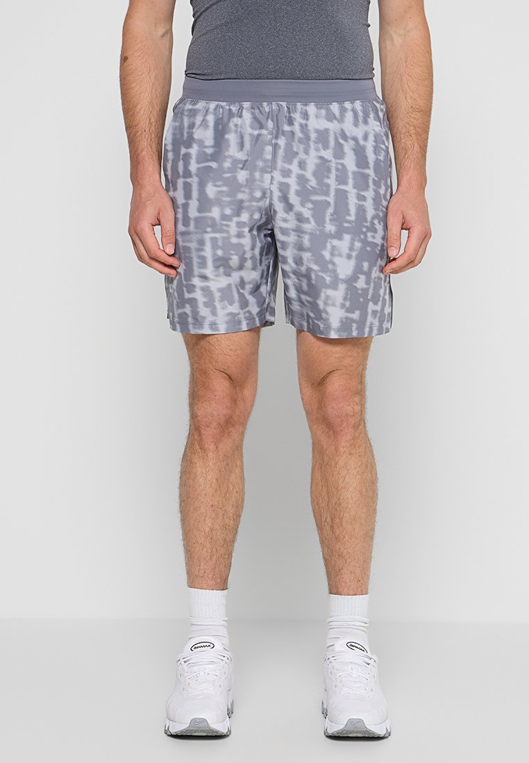 Homme portant un short de sport gris à motifs, un t-shirt gris ajusté, des chaussettes blanches et des baskets blanches, debout devant un fond uni.