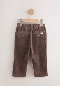 Pantalon en velours côtelé marron pour tout-petit avec taille élastique, poches arrière à boutons et imprimés discrets de visage d'ours, suspendu sur un cintre en bois.