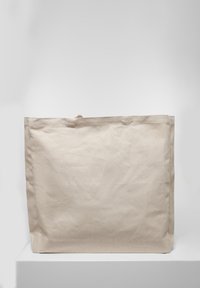 Sac fourre-tout en toile beige uni, debout sur une surface blanche contre un fond gris clair.