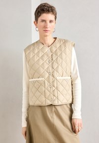 Colete bege acolchoado com fechos de pressão prateados, apresentando dois bolsos frontais e decote redondo, usado sobre uma camisa branca de mangas compridas.