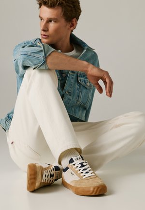 Joven sentado en el suelo, con pantalones blancos, chaqueta vaquera, zapatillas beige y blancas, mirando hacia su izquierda contra un fondo liso.