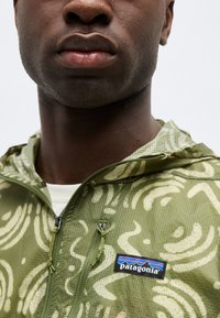 Κοντινό πλάνο ενός ατόμου που φοράει πράσινο μπουφάν Patagonia με ελαφριά αφηρημένα μοτίβα και φερμουάρ στο μπροστινό μέρος σε λευκό φόντο.