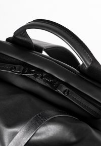 Zaino nero con un design elegante, texture liscia, doppie cerniere e una maniglia in tessuto, che enfatizza uno stile minimalista e la funzionalità.