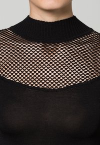 Schwarzes Oberteil mit hohem Kragen, das ein Fischernetzmuster über dem oberen Brustbereich und den Schultern sowie einen einfarbigen schwarzen Strickstoff darunter besitzt.
