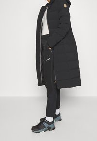 Musta pehmustettu pitkä takki hupulla, jossa on logopatch, vetoketjut sivutaskuissa, yhdistetty mustiin housuihin ja musta-sinisiin tenniksiin.