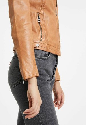 Hellbeige Leder-Zip-Jacke mit tailliertem Schnitt, silbernen Beschlägen und einer horizontalen Reißverschlusstasche. Kombiniert mit dunkelgrauen Jeans.