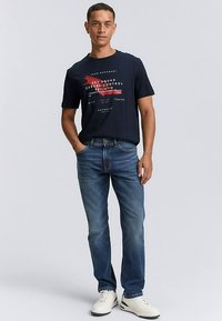 Laivastonsininen puuvillainen t-paita, jossa valkoista ja punaista graafista tekstiä, yhdistetty vaaleansinisiin farkkuihin ja valkoisiin tennareihin. Malli seisoo neutraalissa asennossa.