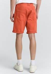 Oransse denim-shortsit käärittyine lahkeineen, joissa on kaksi takataskua ja vyötärönauhan etiketti, yhdistettyinä valkoisiin tennareihin ja ribatuille sukille.