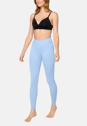 Frau trägt einen schwarzen BH und hellblaue Leggings mit hohem Bund, steht barfuß vor einem schlichten Hintergrund.