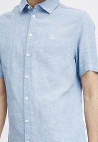 Camicia blu chiaro a maniche corte con bottoni, realizzata in un tessuto testurizzato, caratterizzata da un piccolo logo ricamato sul lato sinistro del petto e bottoni bianchi.