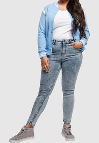 Kvinna iklädd en ljusblå bomberjacka, vit t-shirt, acid-wash skinny jeans och grå höga sneakers, står med en hand i fickan.