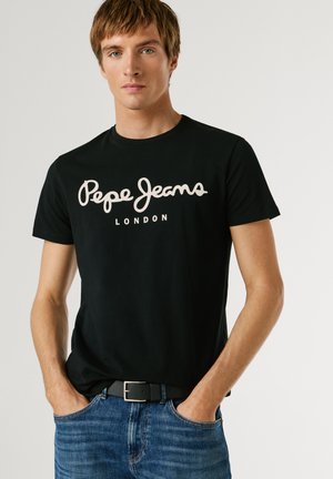Ung mand med hænderne i lommerne iført sort "Pepe Jeans London" T-shirt og blå jeans mod en ensfarvet lys baggrund.