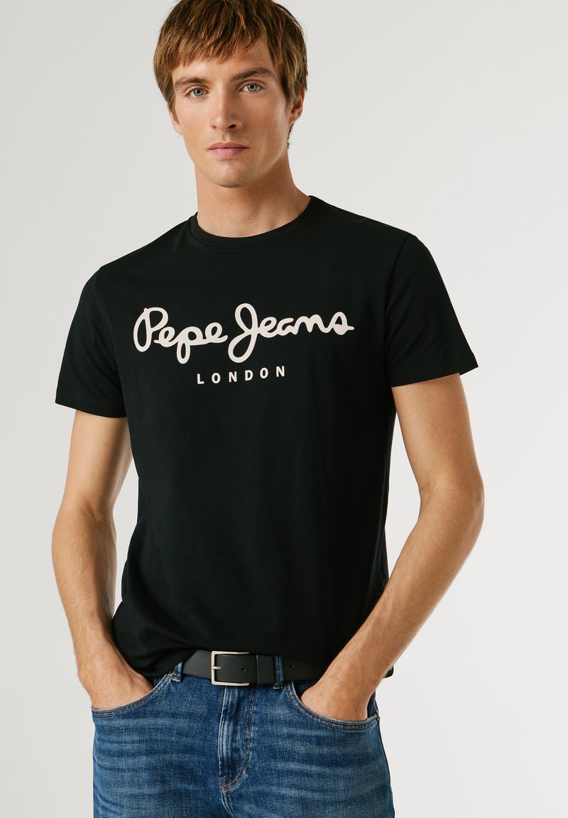 Jeune homme les mains dans les poches portant un T-shirt noir "Pepe Jeans London" et un jean bleu sur un fond clair uni.