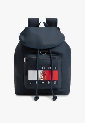Granatowy plecak z klapą, przednią kieszenią na zamek, paskami na ramiona oraz łatką z logo Tommy Jeans, przedstawiającą biały, czerwony i herbowy wzór.