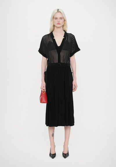 Vestido midi negro con plisados, con una superposición de rayas verticales transparentes, cuello abotonado y un cinturón. Combinado con un bolso rojo y tacones negros.