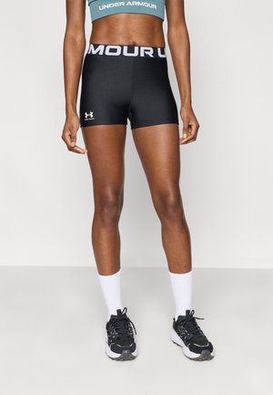Osoba w dobrej formie, ubrana w czarne spodenki Under Armour, białe skarpetki, czarno-białe buty do biegania oraz turkusowy sportowy biustonosz Under Armour.