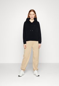 Felpa nera con tasca frontale e cordino, abbinata a pantaloni della tuta beige, completata da sneakers bianche, tessuto morbido, vestibilità rilassata.