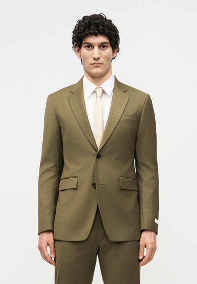 Άνδρας με σγουρά σκούρα μαλλιά που φοράει ελαιδί κοστούμι, λευκό πουκάμισο και κρεμ γραβάτα, στέκεται μπροστά σε απλό φωτεινό φόντο.