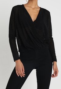 Top nero a maniche lunghe con scollo a V profondo, caratterizzato da un motivo a pois testurizzato e un design arricciato in vita.