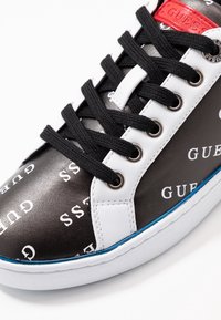 Baskets noir et blanc avec des imprimés du logo "GUESS", dotées de lacets noirs, d'œillets en argent et d'un accent rouge sur la languette. Contour bleu autour de la semelle.