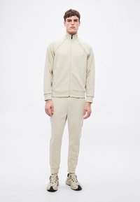 Beżowa kurtka dresowa i dopasowane spodnie z miękkiego materiału. Kurtka ma ściągacze na mankietach i wysoki kołnierz. Do kompletu jasne sneakersy.