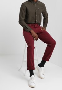 Olijfgroen button-up shirt met een borstzak, gecombineerd met tappende bordeauxrode broek en witte sneakers. Glad stof, opgerolde mouwen en hoog contrast kleuren.