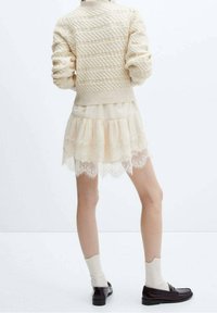 Crème kabelgebreide trui over een beige jurk met kantafwerking. De outfit is voorzien van enkelsokken en donkere instappers, gezien vanuit de achterkant.