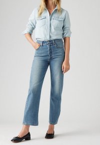 Camisa de denim azul claro con dos bolsillos en el pecho, pantalones vaqueros azules de tiro alto con un corte recto, combinados con zapatos negros de tacón bajo.