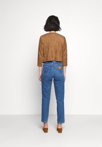 Veste courte en daim marron à manches trois-quarts, associée à un jean bleu coupe droite avec un motif sur la poche arrière. Chaussures marron à talons blocs.