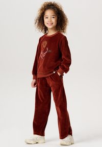 Rostfärgad velour-träningsoverall med rund halsringning, prydd med dekorativa broderade mönster och elastiska ärmavslut, i kombination med lätta sneakers.