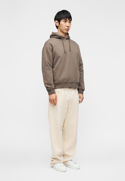 Καφέ φούτερ με κουκούλα και τσέπη καγκουρό, σε συνδυασμό με χαλαρά κρεμ παντελόνια και λευκά αθλητικά παπούτσια, στέκεται μπροστά σε ένα απλό φόντο.