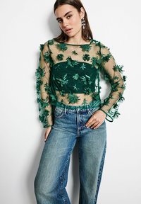 Grün durchsichtige Bluse mit floralen Applikationen, langen Ärmeln und einem quadratischen Ausschnitt, kombiniert mit blauen hochgeschnittenen Jeans.