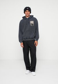 Hanorac gri din bumbac, cu un buzunar frontal și un design grafic, asortat cu pantaloni cargo negri și adidași albi. Pe cap se află o căciulă tricotată neagră.