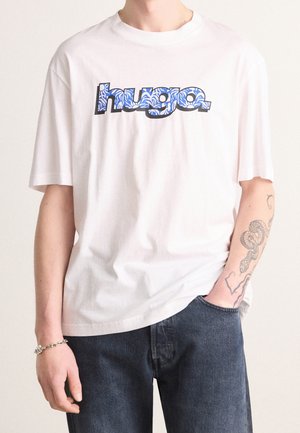Biała bawełniana koszulka z niebieskim wzorzystym logo "Hugo". Krótkie rękawy, luźny krój, noszona razem z ciemnymi dżinsami, z widocznym tatuażem na ramieniu.