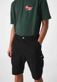Pantaloni cargo neri con tasche laterali, realizzati in tessuto di cotone. Abbinati a una maglietta verde con stampa di testo rossa. Design semplice e funzionale.
