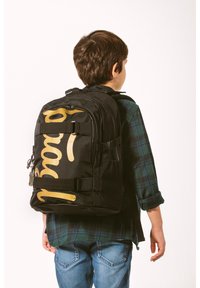 Zaino nero con logo dorato, realizzato in materiale resistente. Dotato di spallacci regolabili, zip e scomparti multipli.
