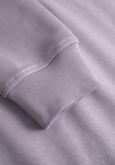 Un pull léger violet en tissu avec une texture lisse et des poignets côtelés, accentué par un design épuré et simple.