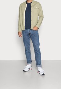 Camisa de botón verde oliva claro sobre una camiseta azul marino, combinada con jeans de mezclilla azules y zapatillas blancas con acentos rojos y marinos.