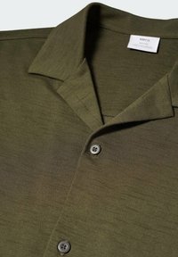 Chemise vert olive à manches courtes avec col camp et deux boutons, montrant un tissu texturé et une étiquette de marque à l'intérieur du col.