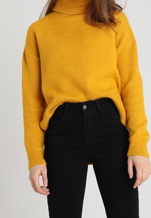 Femme portant un pull à col roulé jaune moutarde rentré dans un jean noir taille haute sur un fond uni.