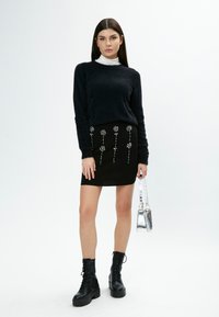 Schwarzer, flauschiger Pullover über einem enganliegenden schwarzen Rock mit floralen Stickereien. Strukturiertes Kampfschuhwerk und eine kleine metallische Handtasche vervollständigen den Look.