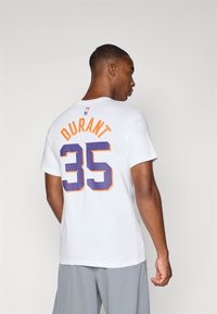 T-shirt bianco con la scritta "DURANT" e il numero "35" in lettere arancioni e viola sul retro. Il tessuto sembra morbido, design casual.