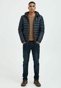 Sort pufferjakke med hætte, struktureret beige sweater, mørke jeans og brune mønstrede sko. Jakken har lynlås og sidelommer.