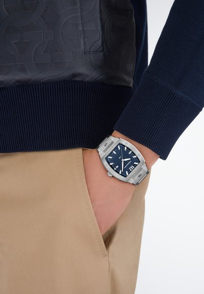 Tengerészkék pulóver bordázott mandzsettákkal, texturált bőrbetéttel. Világos bézs nadrággal és ezüstszínű órával viselve, amelynek kék számlapja van.