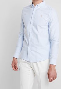Camisa de manga larga azul claro con cuello clásico, que presenta un pequeño logo en el pecho y botones en la parte delantera.